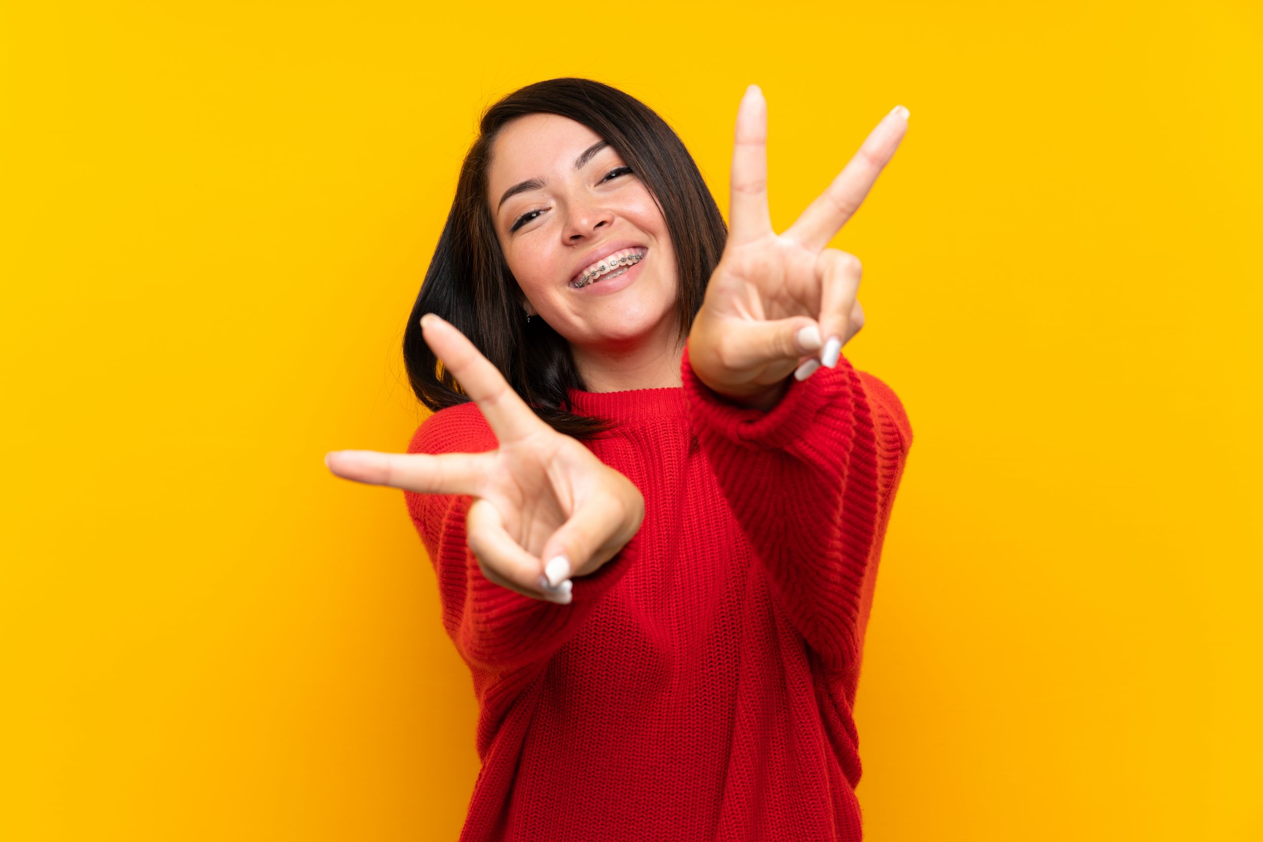 Young Mexican woman with red sweater over yellow wall smiling and showing victory sign