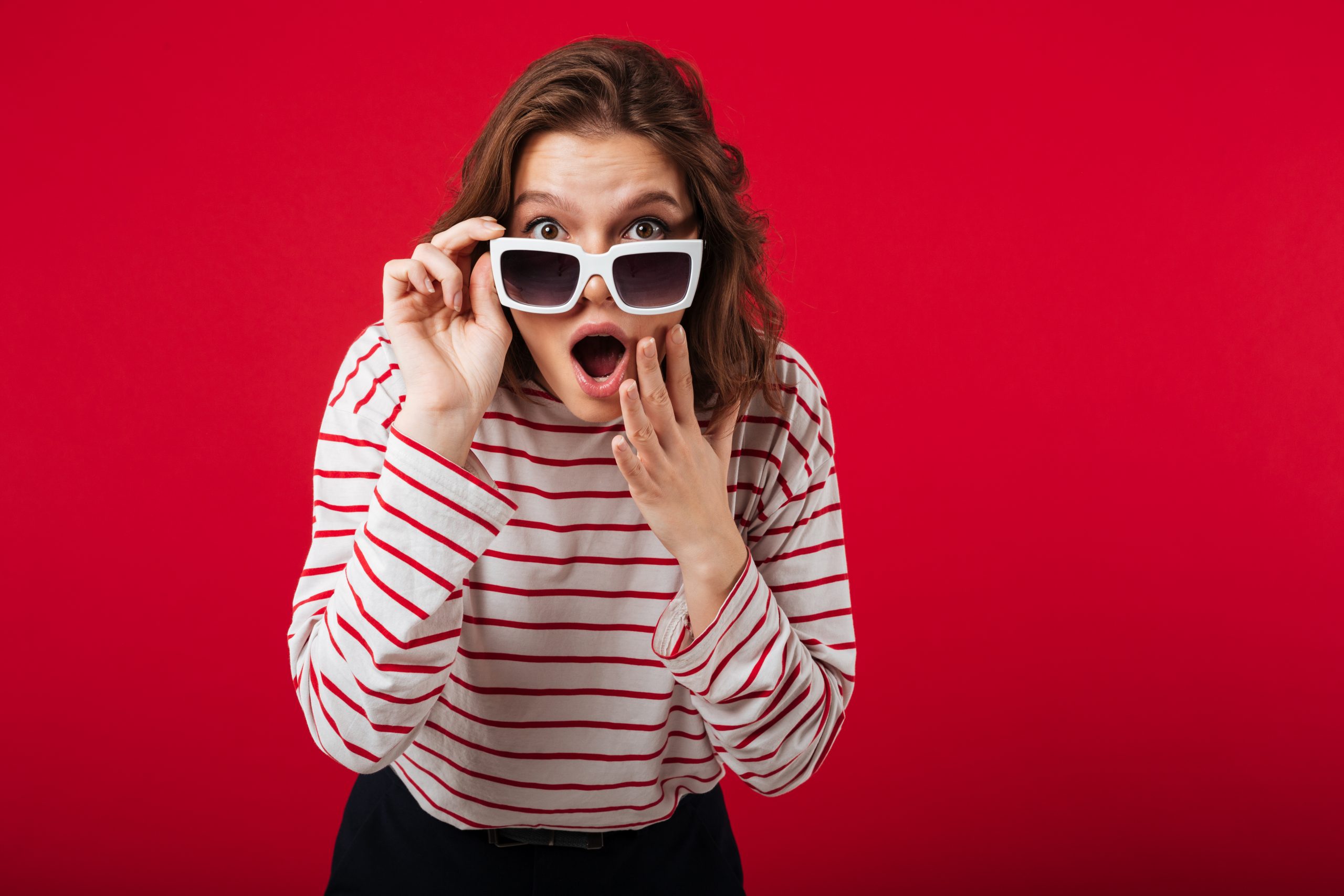 Portrait of a shocked woman in sunglasses posing while standing and looking at camera isolated over pink background