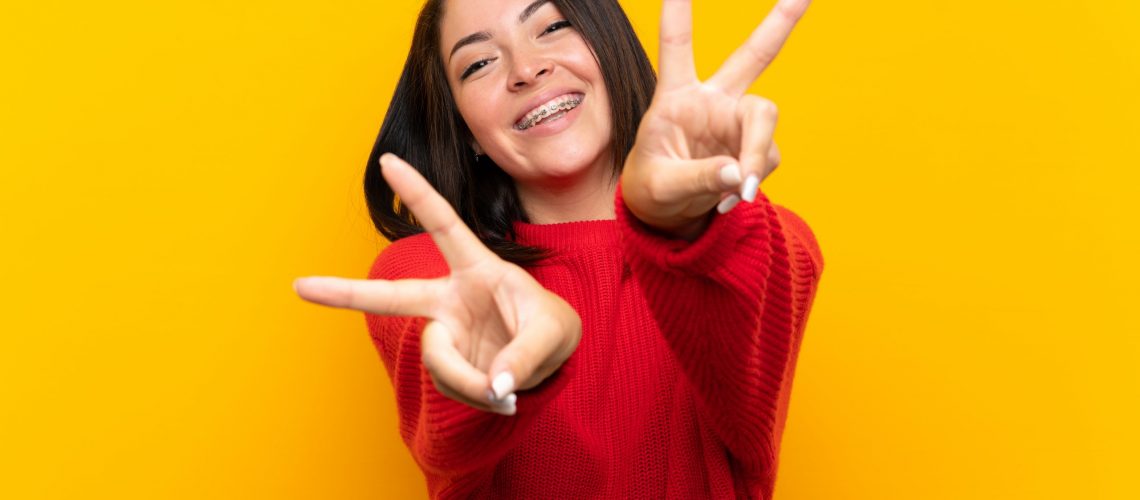 Young Mexican woman with red sweater over yellow wall smiling and showing victory sign