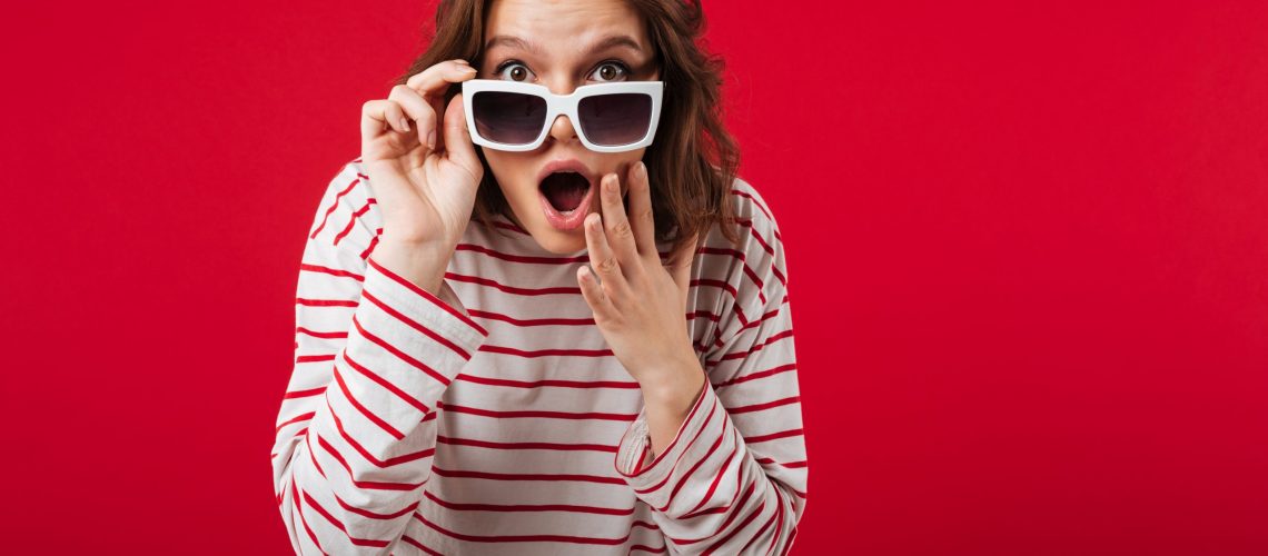 Portrait of a shocked woman in sunglasses posing while standing and looking at camera isolated over pink background