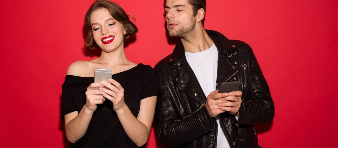 Smiling female punk using smartphone while man peeps in him over red background